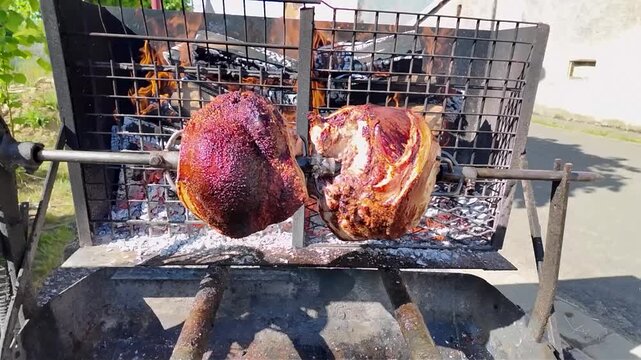 Whole pork shanks roasting slowly on a rotating spit outdoors, over glowing coals. Large pieces of pork rotate slowly on a metal spit over glowing embers and ashes, giving off a delicious aroma. 