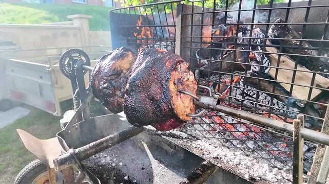 Whole pork shanks roasting slowly on a rotating spit outdoors, over glowing coals. Large pieces of pork rotate slowly on a metal spit over glowing embers and ashes, giving off a delicious aroma. 