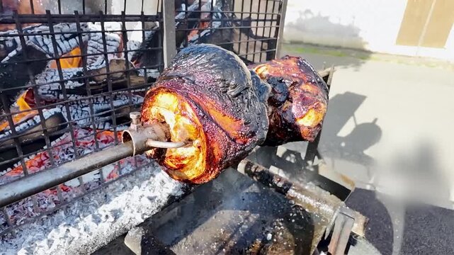 Whole pork shanks roasting slowly on a rotating spit outdoors, over glowing coals. Large pieces of pork rotate slowly on a metal spit over glowing embers and ashes, giving off a delicious aroma. 
