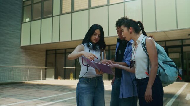 Three high school students collaborate on their studies outside a modern school building. Young friends discuss schoolwork together outdoors.
