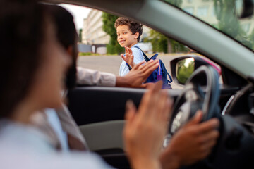 Smiling schoolboy waving goodbye to parents from outside car before heading school