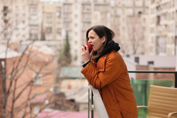 Fototapeta premium Woman in orange quilted coat and black hoodie bites red apple while leaning on balcony railing, with softly blurred apartment buildings behind her and gentle natural light creating relaxed urban