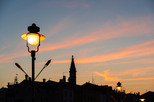 Silhouette of rooftops and the bell tower of Rovinj in Croatia, dominated by a lit street lamp in the foreground.
