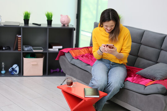 Young woman in yellow hoodie sits on gray sofa and looks at smartphone in bright modern living room, with colorful blanket, red side table, notebook, shelves, and soft daylight around her