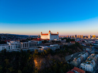 Bratislava castle stands on a hill overlooking the city as the sun sets in the background with buildings and hills around © DSMT