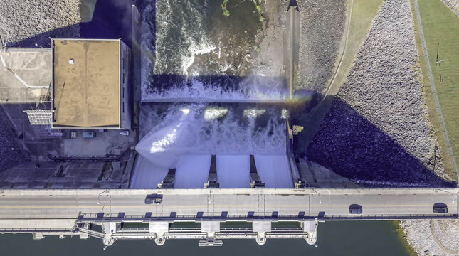 Aerial view of a hydroelectric dam with water rushing through open spillway gates into a basin, with vehicles driving on the road above in Los Angeles, California, United States.
