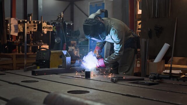 Professional welder wearing protective helmet and gloves welding steel components on workbench in workshop. Clip is suitable for industrial manufacturing and vocational education campaigns