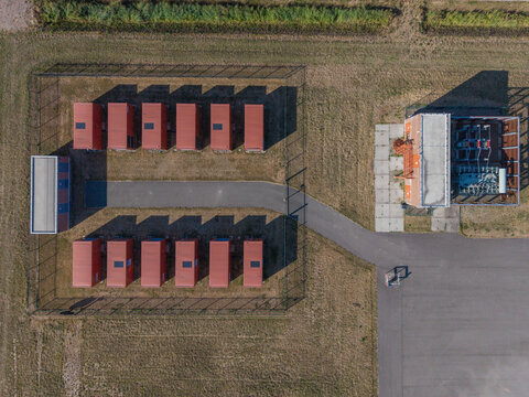 Aerial view of a fenced utility site featuring rows of small red-roofed containers and a transformer station on a grassy field in the Netherlands.