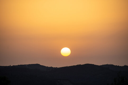 A clear, bright sun with a visible sunspot sets over a dark silhouetted horizon. The vibrant golden-orange sky creates a minimalist and powerful landscape scene