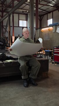 Vertical video of young adult Caucasian male welder sitting in workshop and studying large paper blueprint. Footage is ideal for industrial manufacturing and engineering concepts