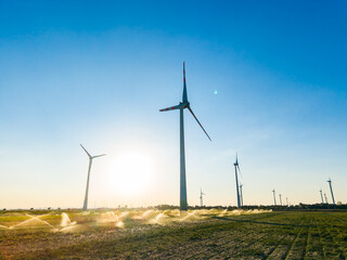 Obraz premium Wind turbines stand tall over fields with sprinklers operating at sunset near a farming area