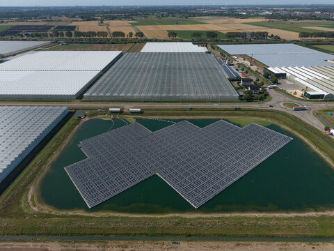 Aerial view of a large floating solar panel array on a pond, situated next to extensive glass greenhouses and agricultural fields in the Netherlands.