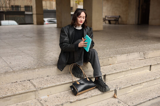 Young woman sits on worn stone steps in city plaza, holding turquoise notebook with black handbag nearby, wearing dark layered streetwear and black boots, with calm reflective expression and soft