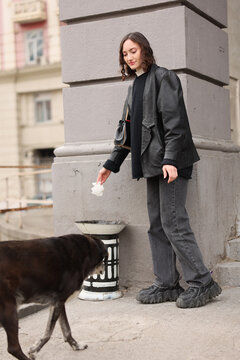 Young woman in dark layered clothing stands beside gray stone wall on city sidewalk, looking down with calm expression while holding tissue near stray dog moving through foreground, creating quiet