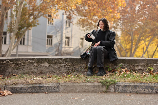 Young woman in dark clothing sits on low stone wall in autumn park setting, holding coffee cup while working on laptop, with fallen leaves, warm colors, soft daylight, and blurred urban buildings