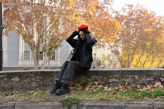Young woman sits on stone wall in urban park during autumn, adjusting red beret and looking down, wearing black jacket, dark jeans and boots, with fallen leaves, warm trees and soft daylight behind