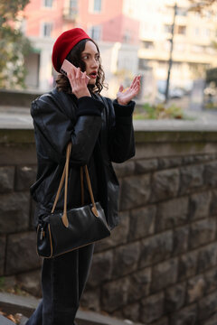 Fashionable woman in black coat and red beret speaks on smartphone outdoors, looking surprised and gesturing with raised hand beside softly blurred urban buildings, stone wall, and natural daylight