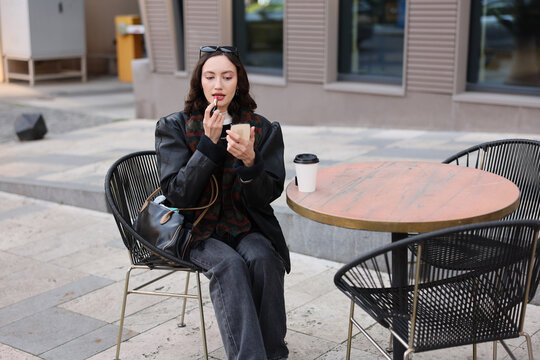 Young woman sits outdoors at cafe table and applies lipstick while holding compact mirror, with handbag and coffee cup nearby, dressed in dark stylish clothing within relaxed city setting
