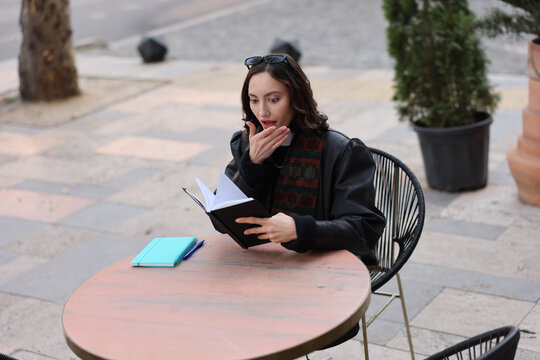 Young woman sitting at outdoor caf table reading open notebook with surprised expression, hand near mouth, pen and second notebook beside her, in soft natural daylight with blurred street background