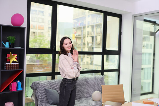 Smiling young woman stands indoors with hands clasped beside sofa and shelving in bright modern apartment, with large windows, soft daylight, colorful decor, and calm relaxed home setting