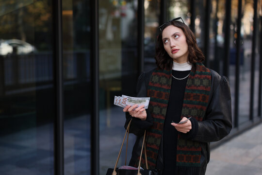 Thoughtful young woman standing beside glass storefront on city sidewalk, holding cash and cards while looking upward, wearing dark outfit with patterned scarf, soft natural light and shallow depth