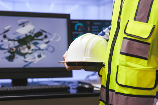 Worker in a safety vest holding a helmet beside a computer screen a drone model in an office setting