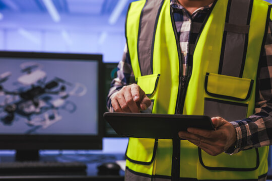 Technician in a safety vest using a tablet in a workshop with a computer monitor in the background