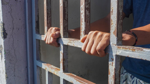 A close-up of an elderly prisoner's hands gripping metal bars.
