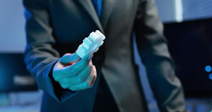 Business person in a dark suit holding a white plastic gear in a modern office, engineering, manufacturing, innovation and teamwork in a blue toned setting.