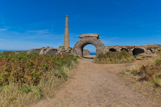 Historic stone ruins under a clear blue sky, ideal for travel content. A tall chimney and a large archway stand on a path leading to the distant sea - Jacob's Mine, Botallack, Cornwall, UK