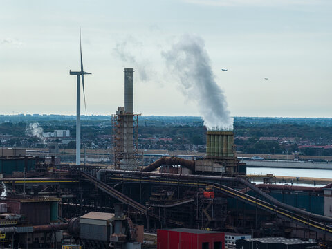 Aerial view of the Tata Steel industrial plant featuring a smoking chimney, a wind turbine, and complex steel infrastructure in Velsen-Noord, Noord-Holland, Netherlands.