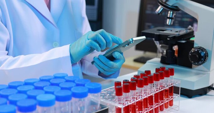 Scientist in a lab coat using a tablet beside a microscope and rows of blood test tubes in a modern laboratory setting.
