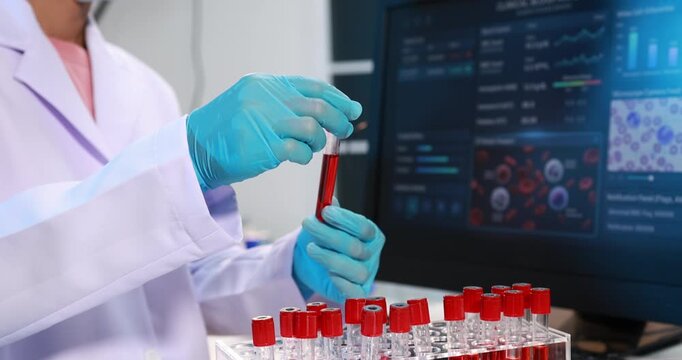 Scientist in a lab coat and gloves holding a blood sample tube near test vials and a medical data screen in a modern laboratory.