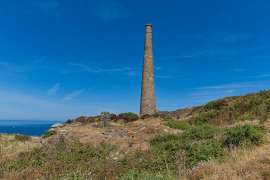 A historic stone chimney stands tall on a grassy cliff overlooking the blue sea under a bright sky - Jacob's Mine, Botallack, Cornwall, UK