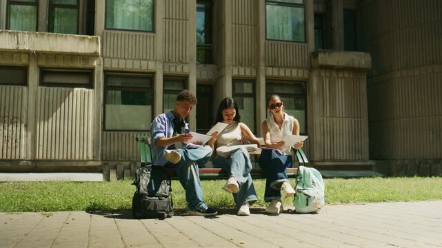 Three high school students sit on a bench outdoors, intently studying and discussing academic papers. Young learners engage in collaborative learning on a sunny day outside a school building.
