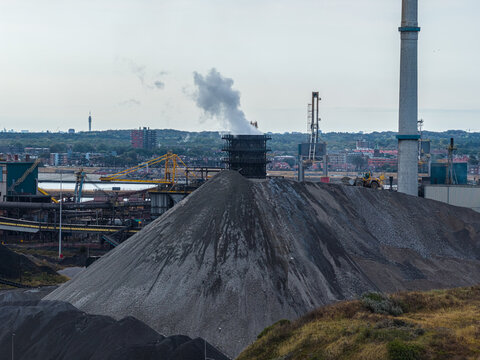 Aerial view of the Tata Steel industrial complex featuring a large pile of raw materials, industrial machinery, and smoke rising from a stack in Velsen-Noord, Noord-Holland, Netherlands.