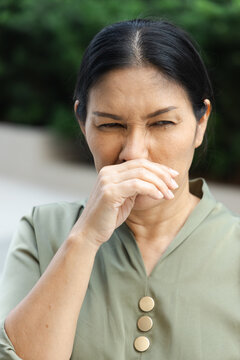 asian middle aged sick woman sneezing with runny nose outdoors in a park, wiping nose with bare hand