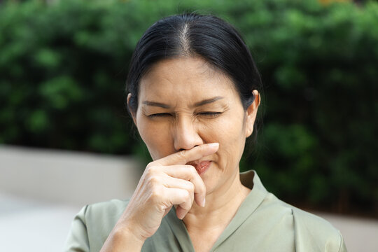 asian middle aged sick woman sneezing with runny nose outdoors in a park, wiping nose with bare hand