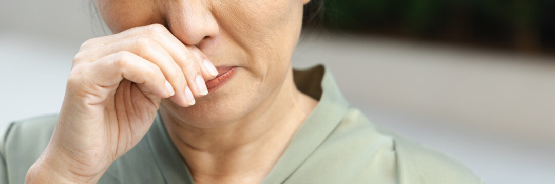asian middle aged sick woman sneezing with runny nose outdoors in a park, wiping nose with bare hand