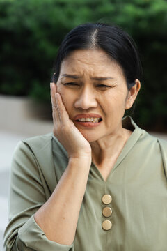 asian middle aged woman suffering from toothache and tooth decay outdoors, holding her cheek in pain, dental care concept, head and shoulder shot