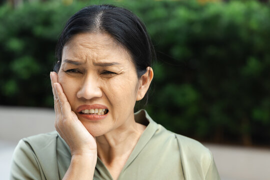 asian middle aged woman suffering from toothache and tooth decay outdoors, holding her cheek in pain, dental care concept, head and shoulder shot