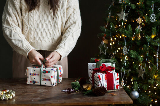 Woman in cozy Knit Sweater Wrapping Festive Presents with Green Ribbon on Rustic Wooden Table near Glowing Tree. Concept of winter DIY, and festive seasonal preparation