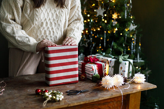 Woman in Cozy Knit Sweater Folding Red and White Striped Wrapping Paper Around a Gift Box on a Wooden Table near festive xmas tree. festive seasonal preparation