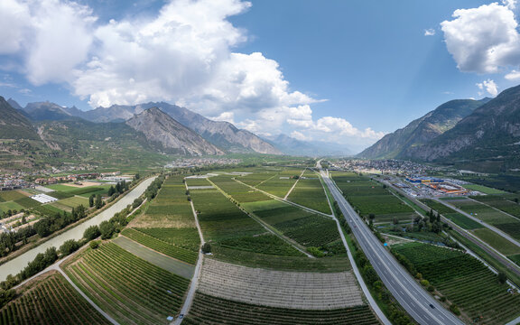 Aerial view of the River Rhone flowing through the valley, flanked by agricultural fields, vineyards, the A9 highway, and an industrial zone in Riddes, Valais, Switzerland.