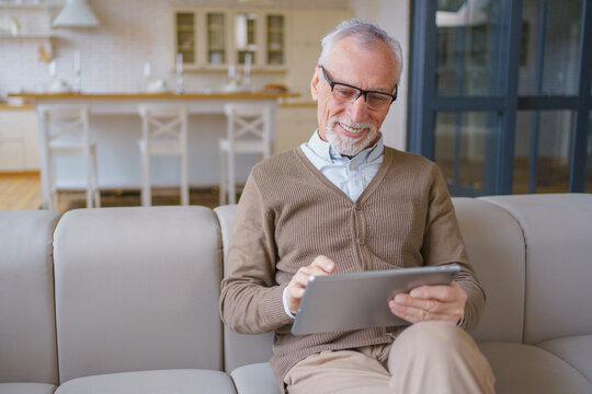 Elderly man sitting on couch and using tablet in living room, senior male enjoying technology at home, browsing internet, relaxed retirement lifestyle and digital communication concept