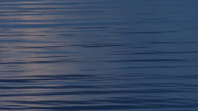 Moonlight reflection on a calm blue sea in the early evening, where gentle waves and ripples create horizontal, parallel lines. Close-Up