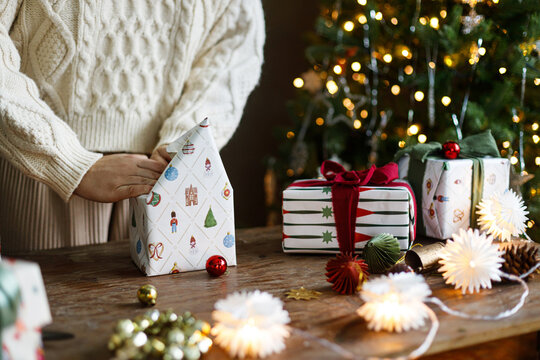 Woman in Cozy Sweater Folding Festive Patterned Wrapping Paper Around a gift Box on Rustic Table. Atmospheric christmas mood. winter home traditions, and Christmas surprises