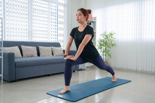 Young Asian woman performing lunge stretch on yoga mat in bright modern living room during home fitness routine