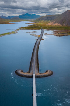 Aerial view of a road stretching across a calm blue lake, featuring a unique T-shaped stone embankment surrounded by mountains under a cloudy sky, ICELAND.