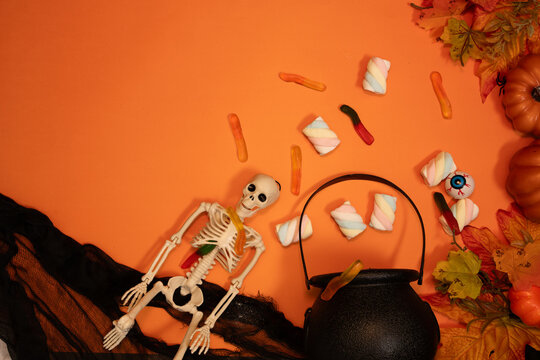 A cheerful Halloween scene shows a skeleton beside a black cauldron filled with colorful candy, pumpkins, autumn leaves, and gauze, creating a playful seasonal arrangement with strong visual appeal.
